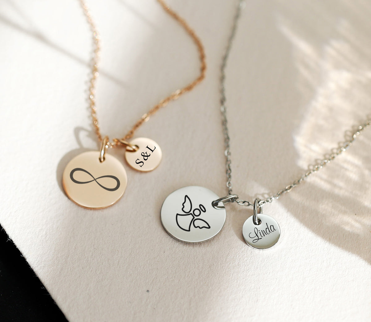 Zwei personalisierte Ketten nebeneinander: Infinity-Plättchen 'S&L' und Engelmotiv mit 'Linda'