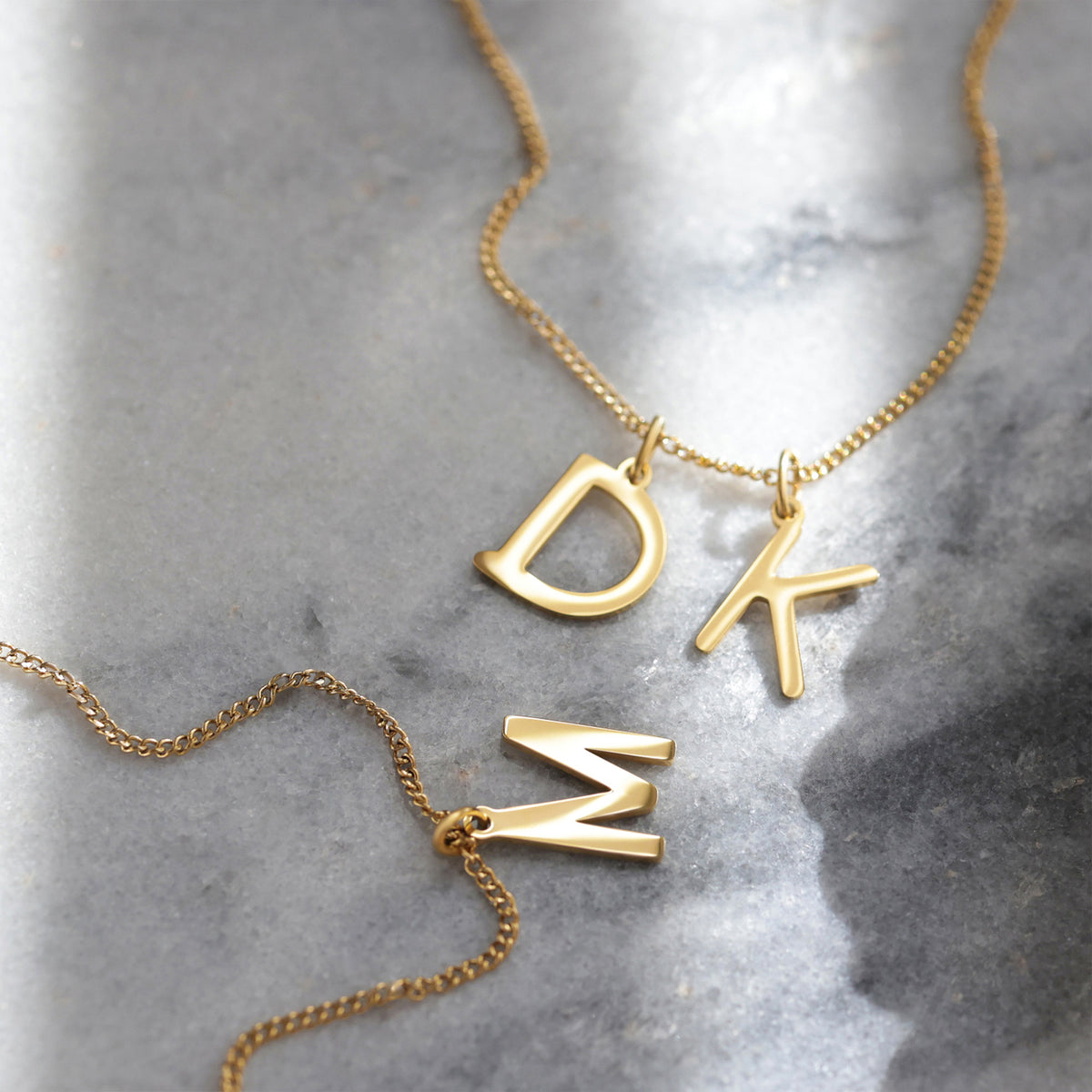 Goldene Ketten mit Buchstaben-Anhängern D, K und M auf Marmoruntergrund