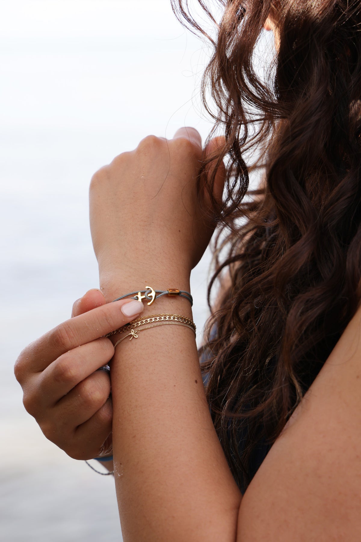 Frau trägt graues Unisex Armband mit Gravur & Ankerhaken mit Bernsteinperle am Strand, kombiniert mit Goldkette
