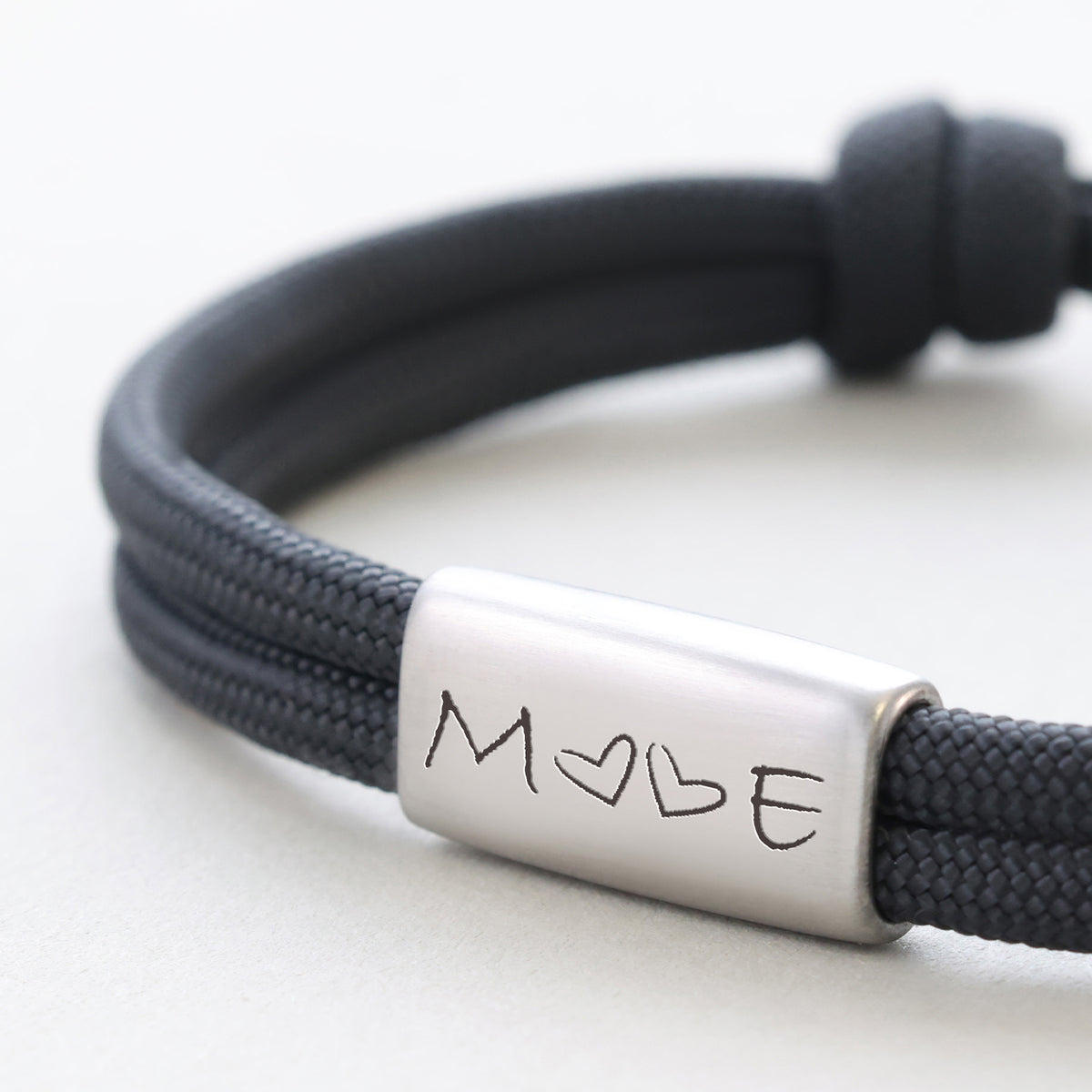 Nahaufnahme eines schwarzen Segeltau-Armbands, graviert mit M und Herz E