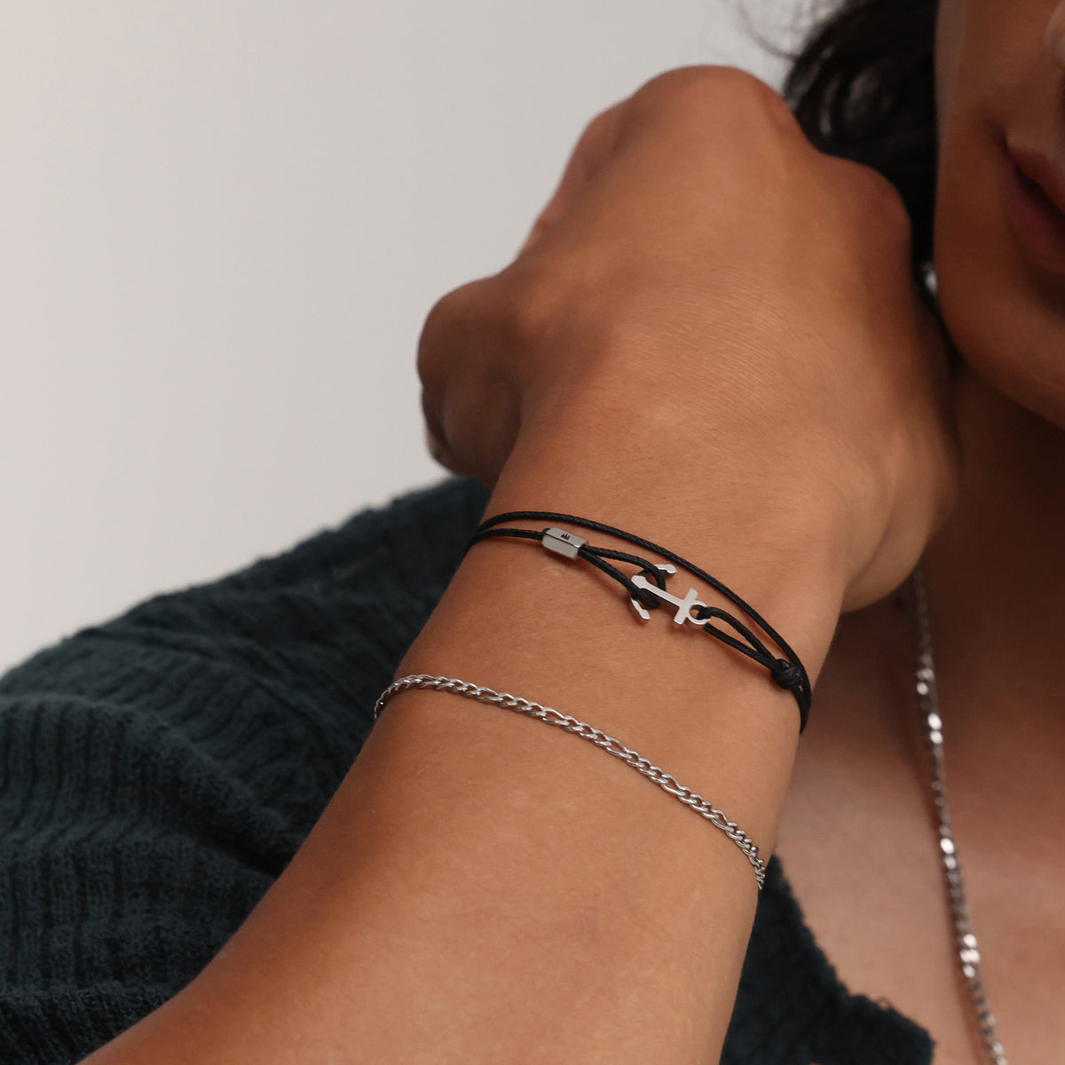 Frau trägt schwarzes Unisex Armband mit Ankerhaken & Gravur kombiniert mit Silberkette am Handgelenk