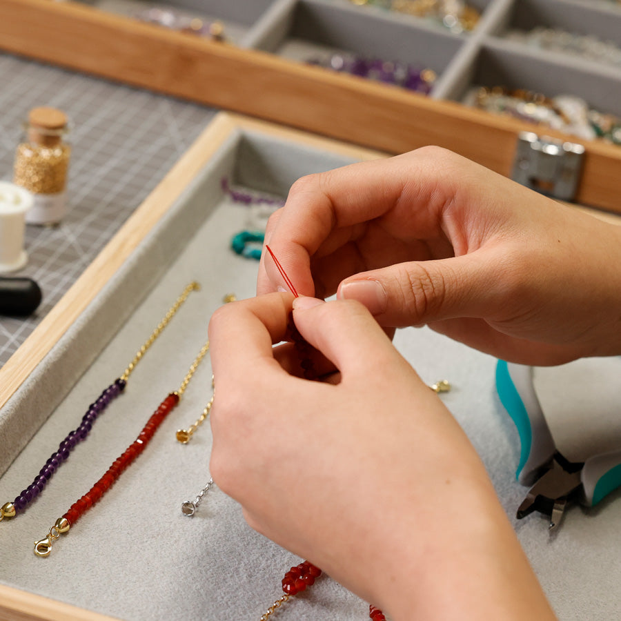 Hände fädeln Edelsteinarmbänder in Rot und Violett an der Werkbank mit Sortierschale