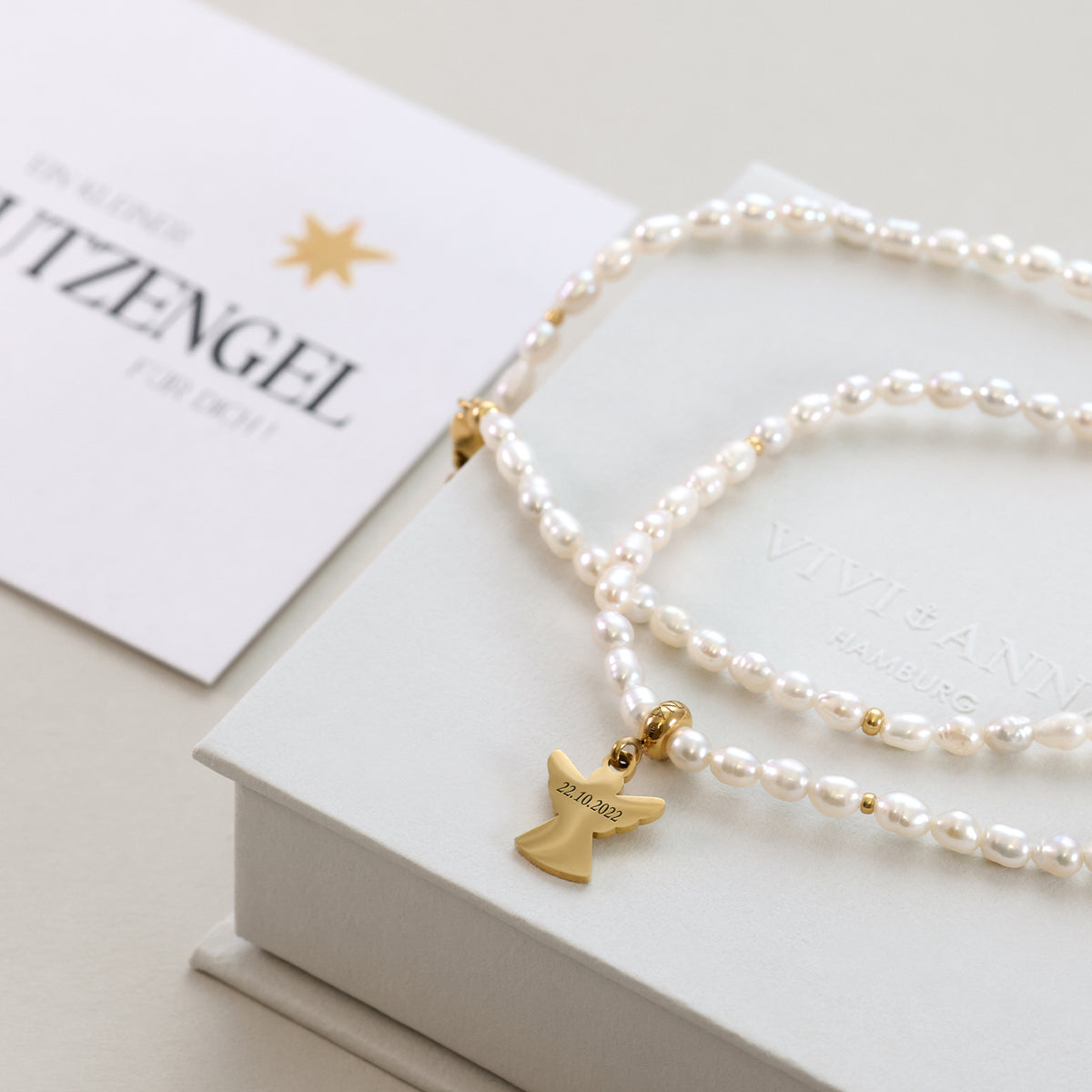 Schutzengel-Perlenkette mit graviertem Datum 22.10.2022, drapiert über Geschenkbox und Karte