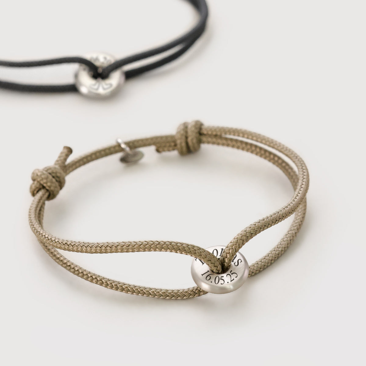 Segeltau Donut Surferarmband in Beige und Schwarz mit silbernem Plättchen im Vergleich