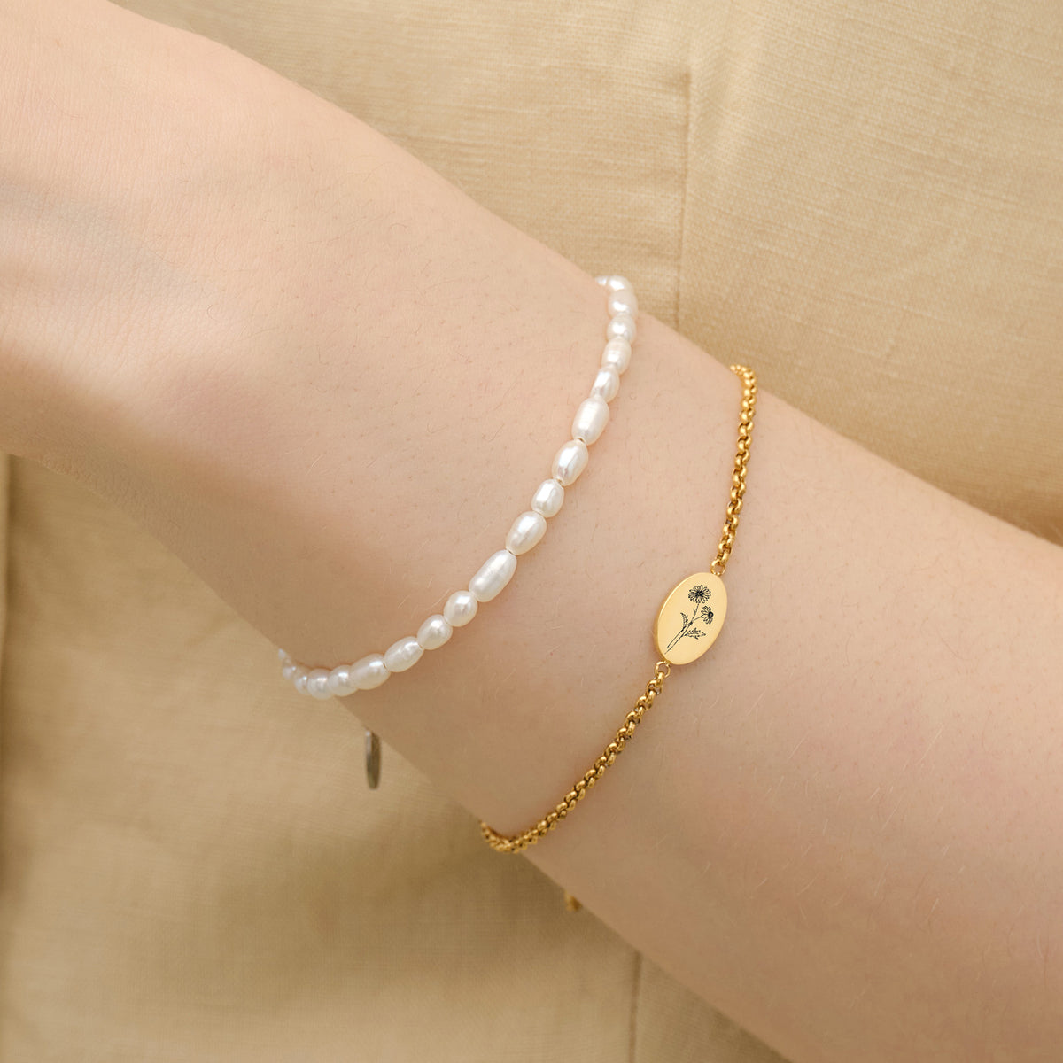 Frau trägt Geburtsblumen-Armband mit ovalem Gravurplättchen in Gold, kombiniert mit Perlenarmband