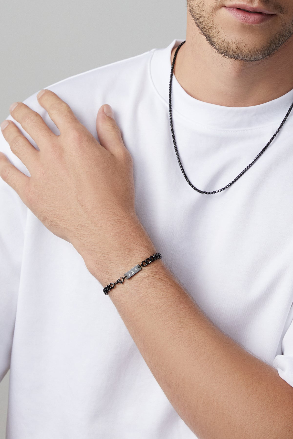 Nahaufnahme: Mann trägt schwarzes Panzerarmband mit Gravurplättchen 'J & A' und passende Halskette