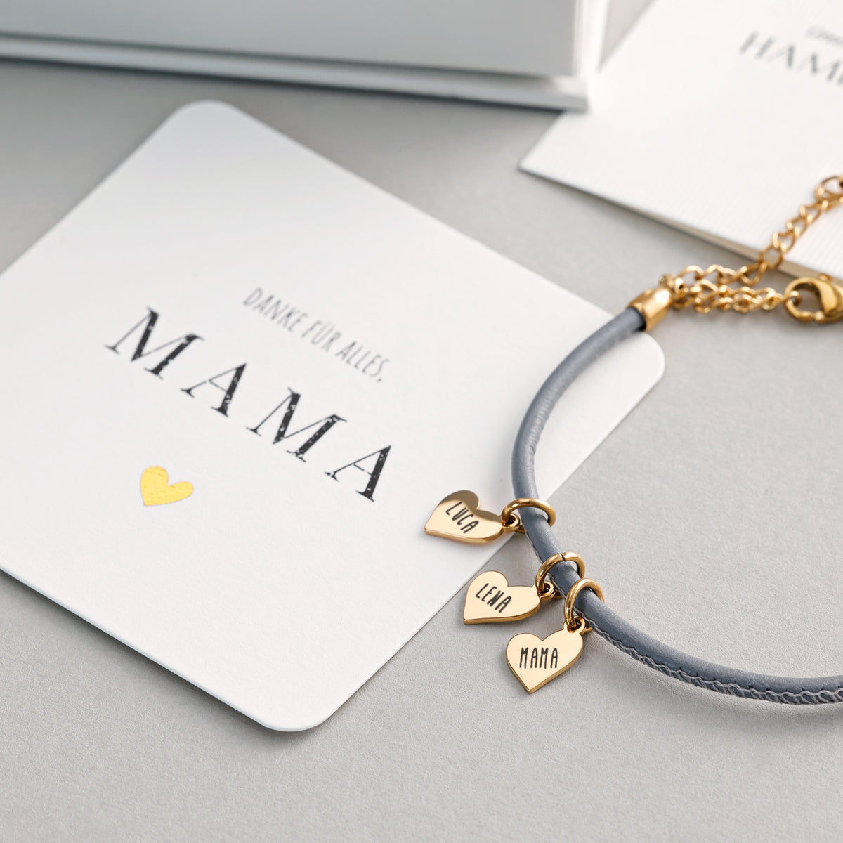 Lederarmband mit Gravur in Grau auf Grußkarte, Herzanhänger graviert mit Luca, Lena und Mama