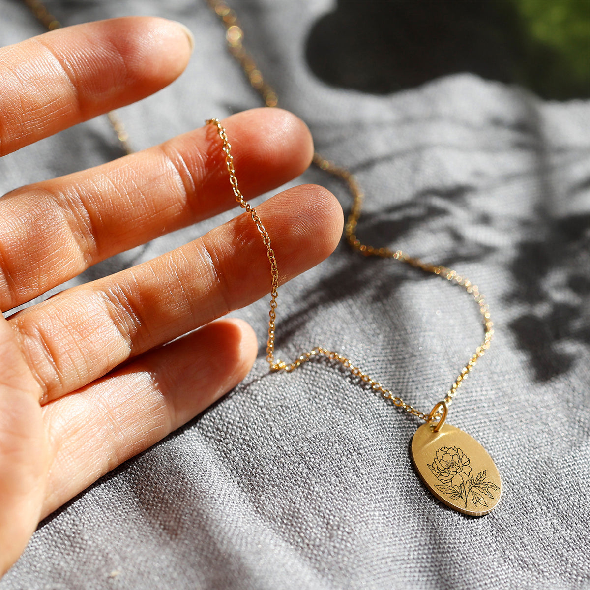 Hand hält goldene Geburtsblumen Kette mit ovalem Anhänger und Pfingstrosen-Gravur im Sonnenlicht