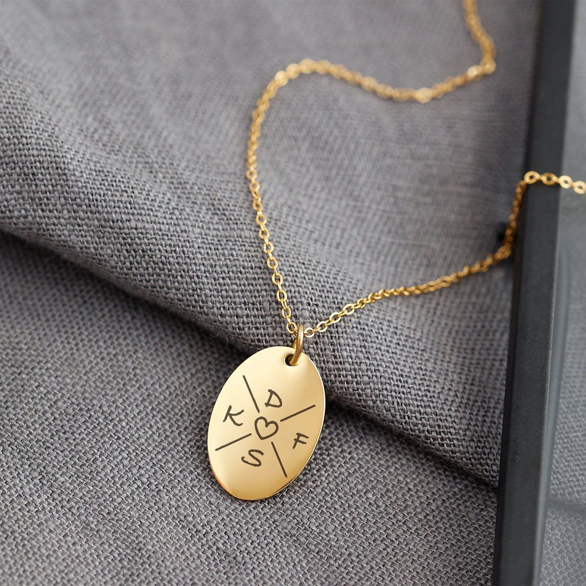 Kette mit graviertem Ovalanhänger in Gold, Initialen und Herzmotiv, auf grauem Stoff