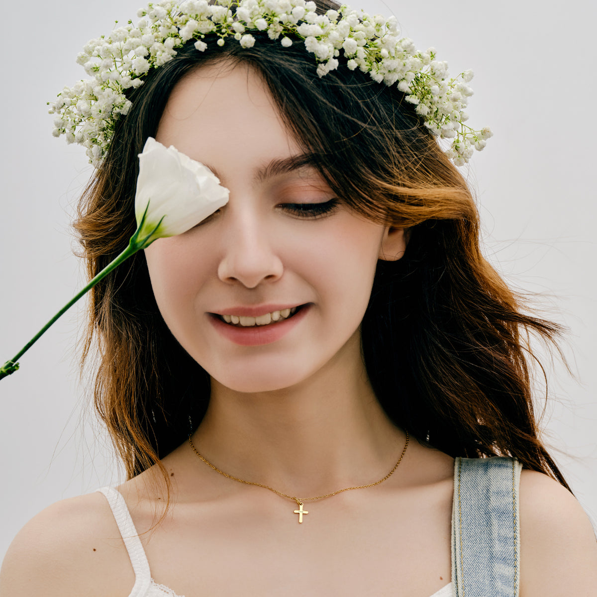 Mädchen mit Blumenkranz trägt Mini-Kreuzkette in Gold, Auge mit weißer Rose verdeckt