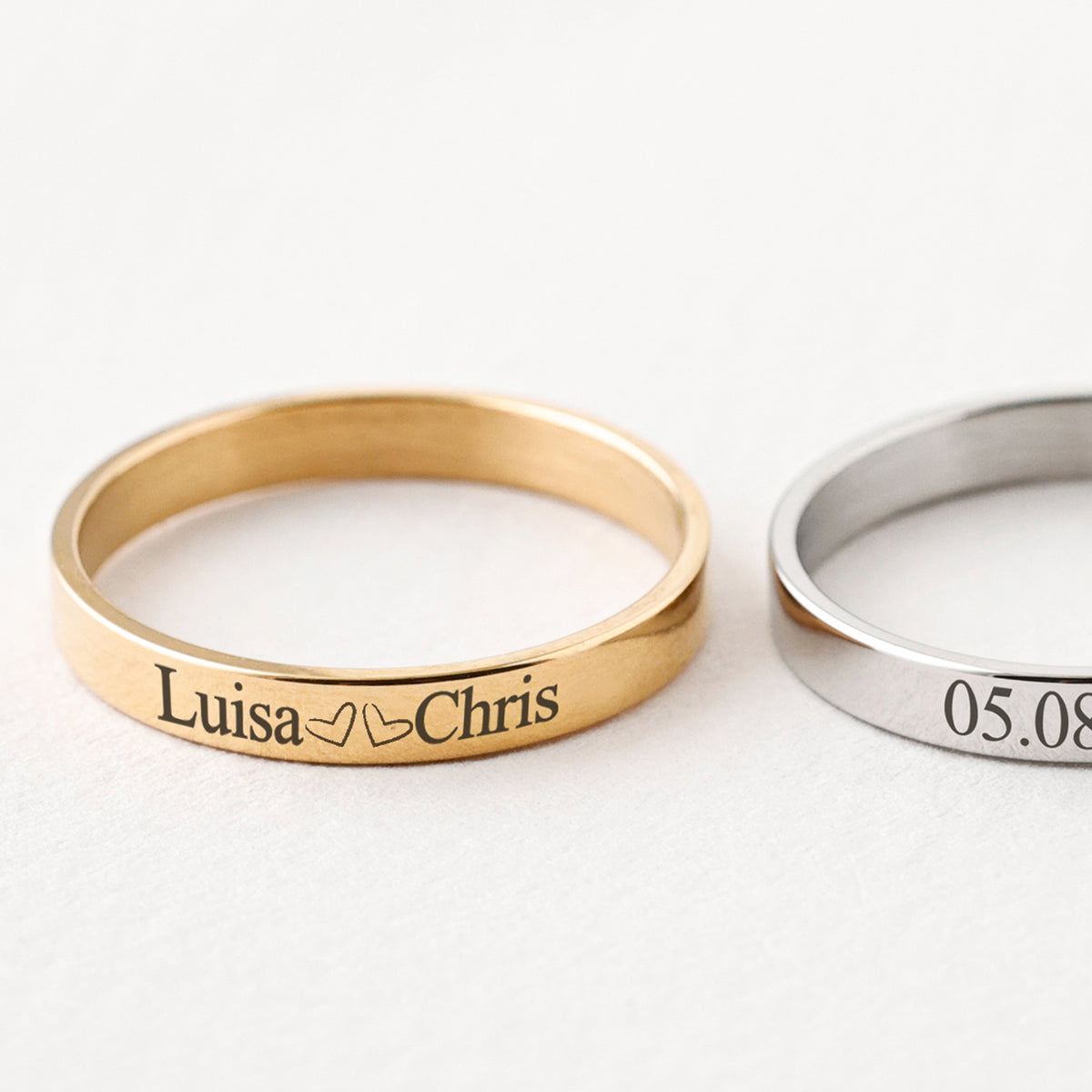 Minimalistischer Gravur-Ring in Gold mit Namen „Luisa & Chris“ neben silbernem Ring mit Datum