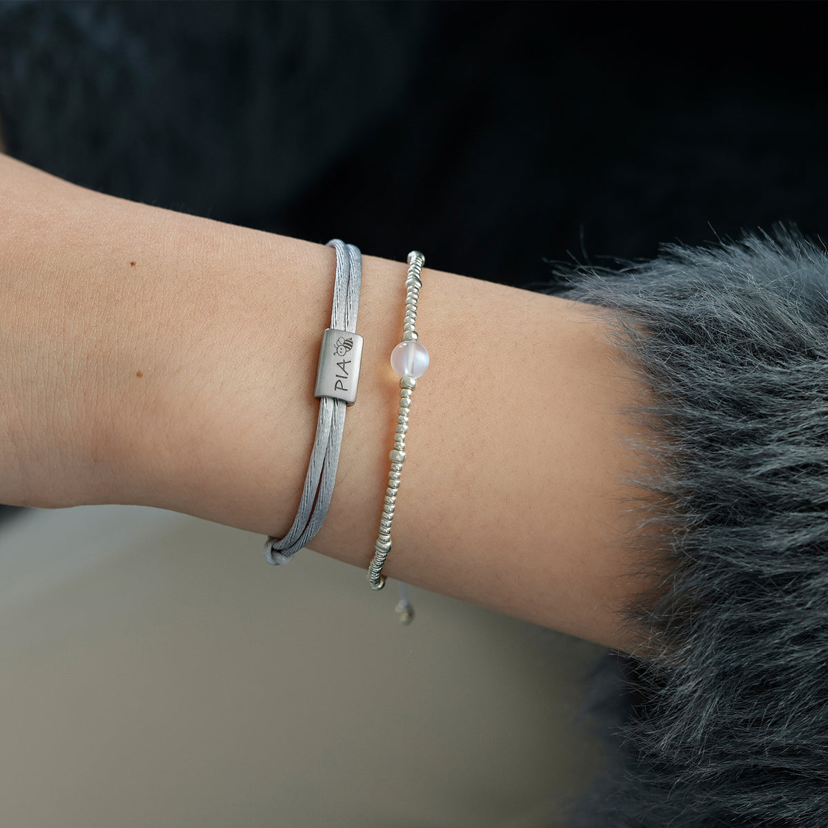 Person trägt graues Kinder Satinband Armband mit Gravur Pia und Pfoten-Symbol, kombiniert mit Perlenarmband