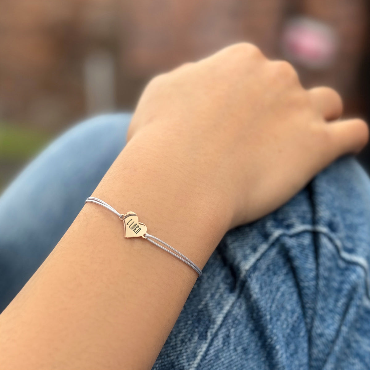 Feines Herz-Armband mit Wunschtext in Grau – Frau trägt Armband mit Gravur 'Clara' auf Jeans