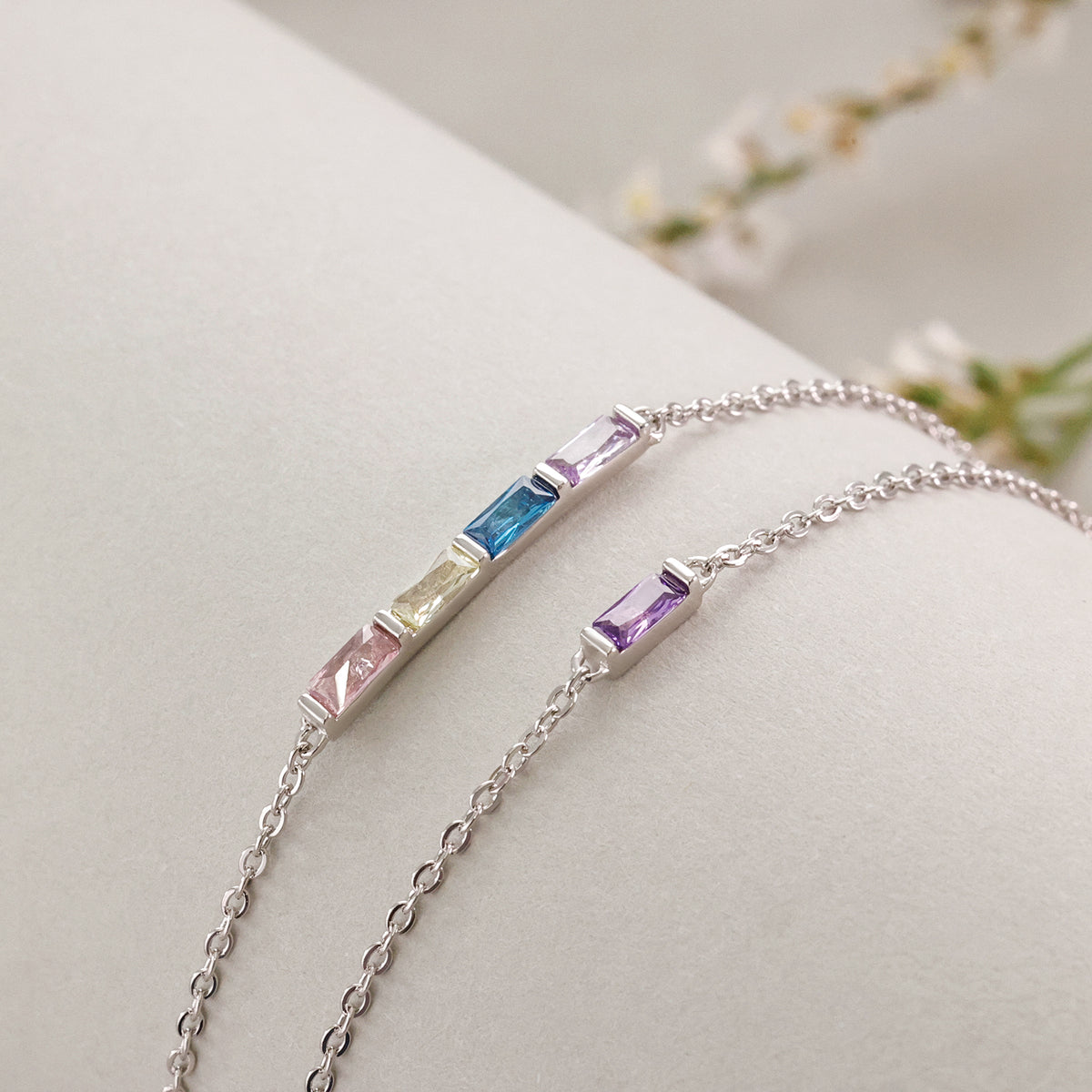 Personalisiertes Geburtsstein-Armband in Silber mit bunten Baguette-Steinen neben weißen Blüten