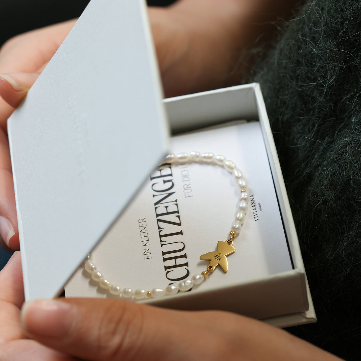 Frau öffnet Geschenkbox mit Perlenarmband und Schutzengel-Anhänger