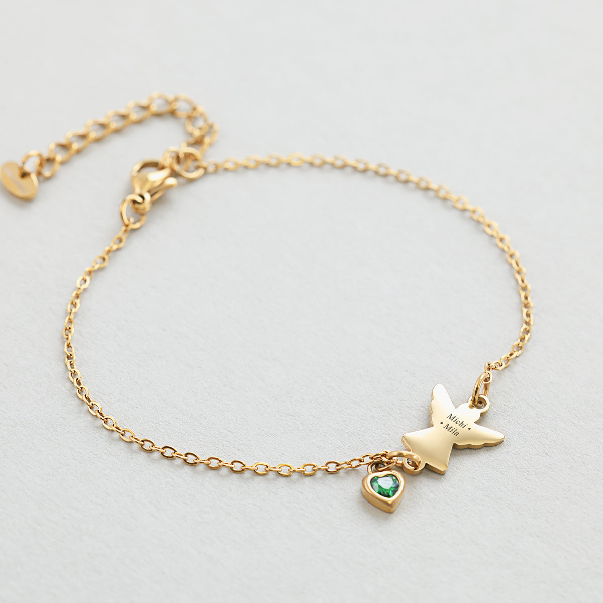 Engel Armband mit Zirkonia Geburtsstein in Gold – Flatlay mit graviertem Engelsanhänger 'Michi Mila' und grünem Stein