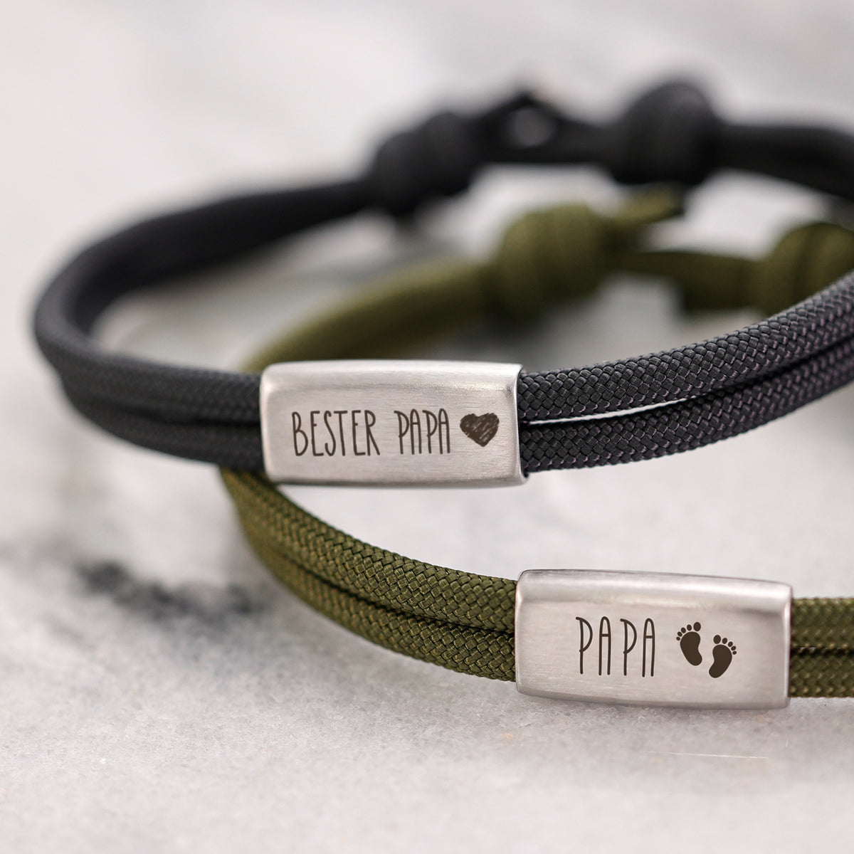 Zwei Papa Armbänder in Schwarz und Oliv – graviert mit 'Bester Papa' und 'Papa' mit Fußabdrücken