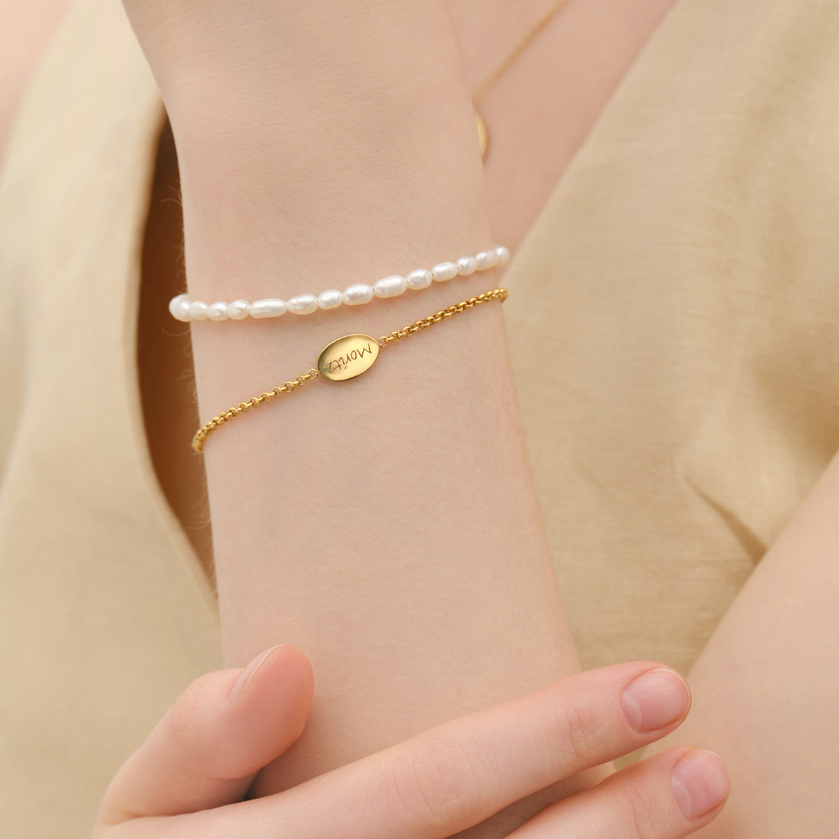 Frau trägt goldenes Armband mit graviertem ovalem Plättchen, kombiniert mit Perlenarmband