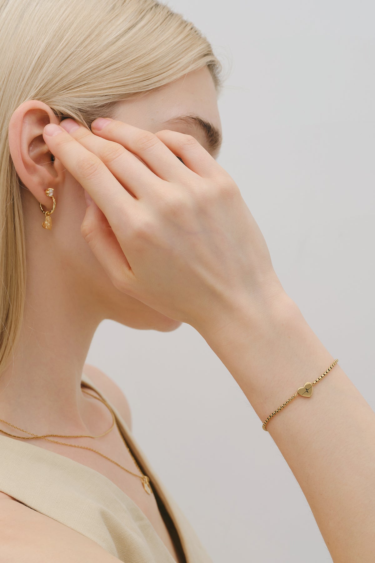 Frau trägt personalisiertes Herz-Armband in Gold am Handgelenk, mit Ohrringen im Hintergrund
