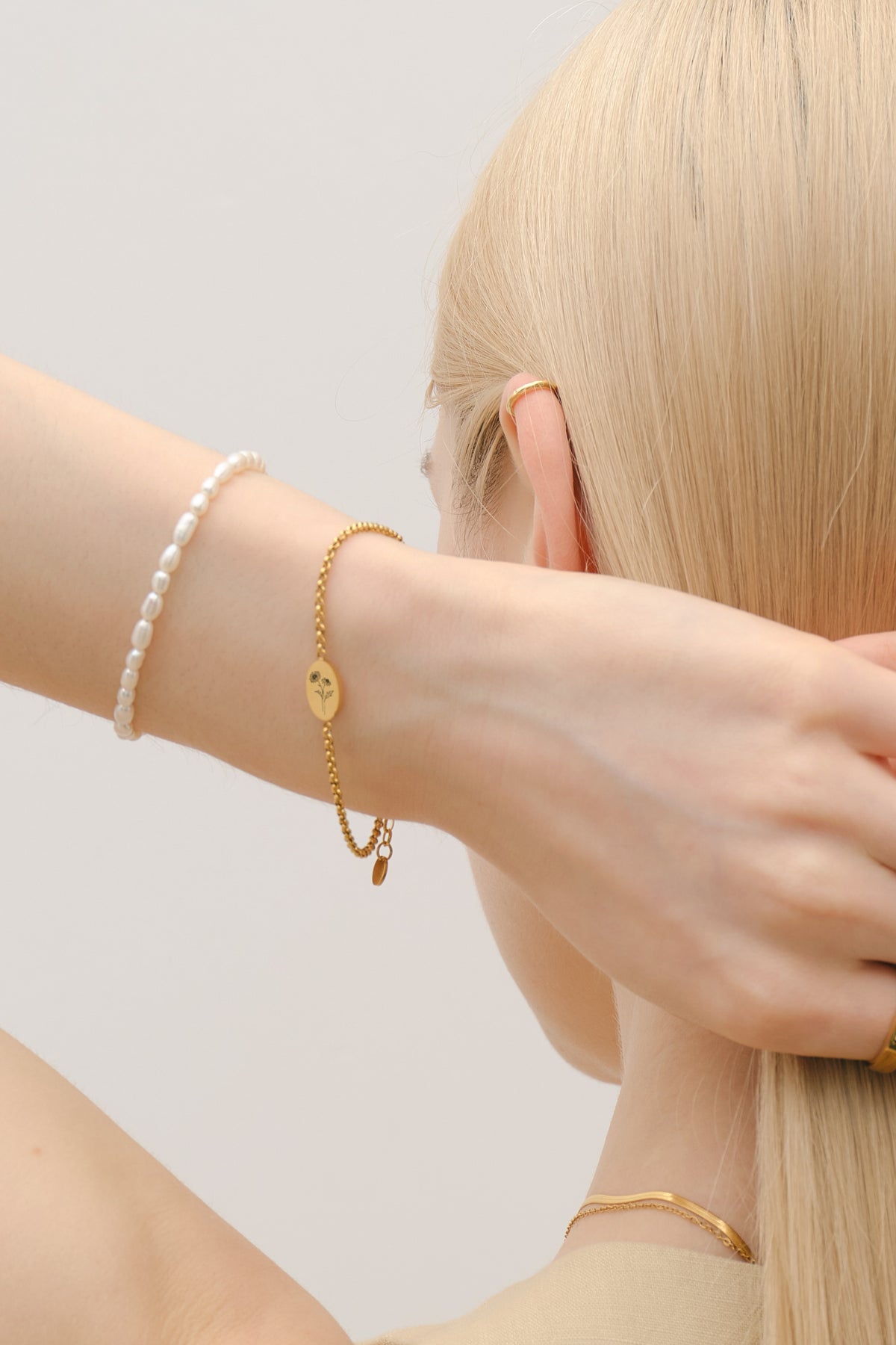 Frau trägt Geburtsblumen-Armband mit ovalem Gravurplättchen in Gold, Rückenansicht mit Perlenarmband