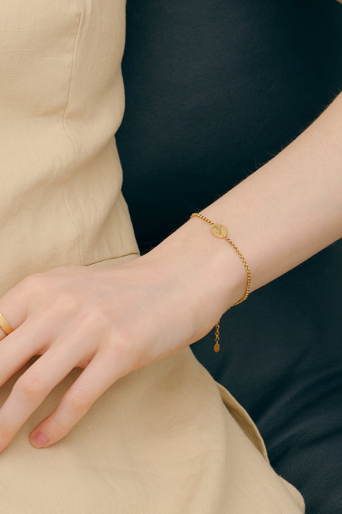 Frau trägt Geburtsblumen-Armband mit ovalem Gravurplättchen in Gold am Handgelenk, sitzend