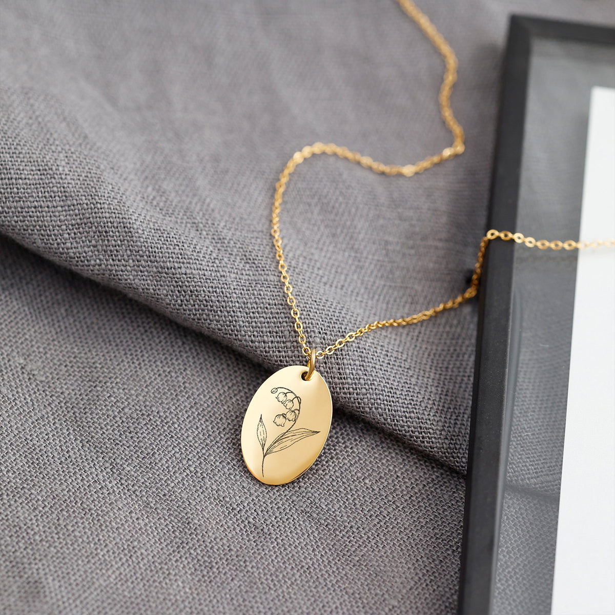 Goldene Geburtsblumen Kette mit ovalem Anhänger – Maiglöckchen-Motiv graviert, auf grauem Stoff