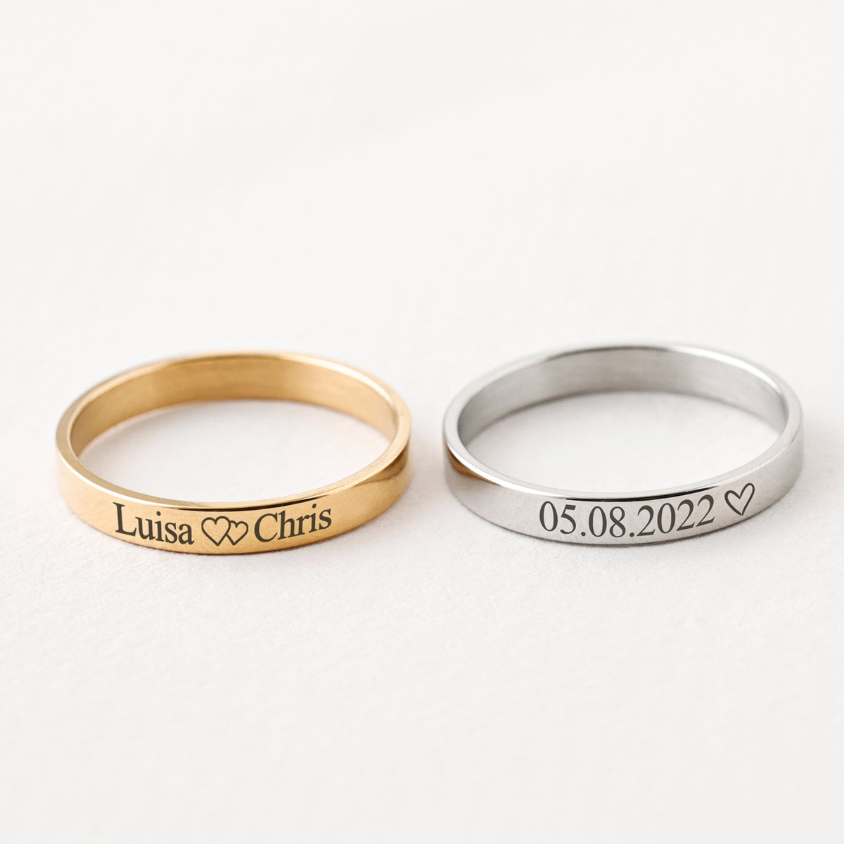 Gravurring in Gold mit Namen 'Luisa Chris' und Herzen neben silbernem Ring mit Datum 05.08.2022
