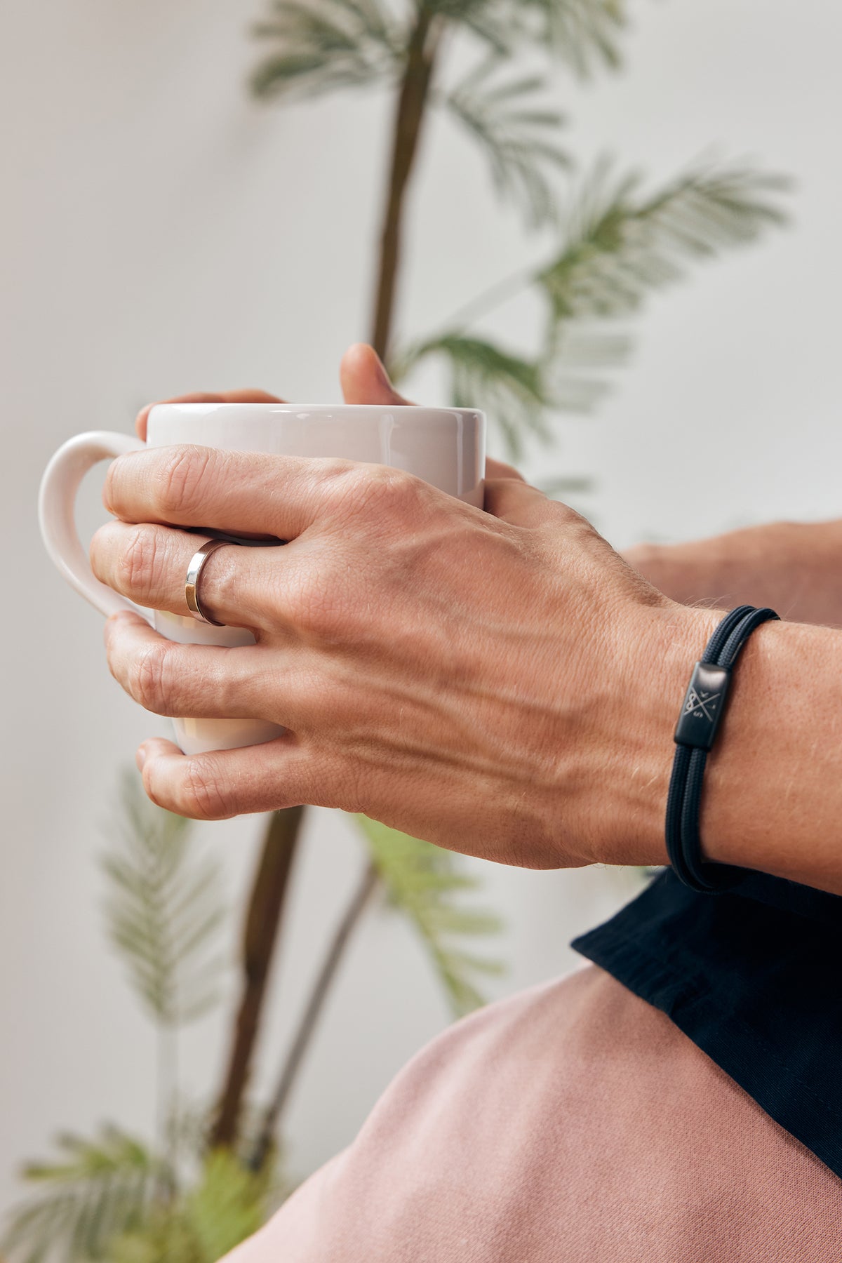 Mann trägt Herren Stapelring mit Gravur in Silber beim Kaffeetrinken, mit Armband und Pflanze im Hintergrund