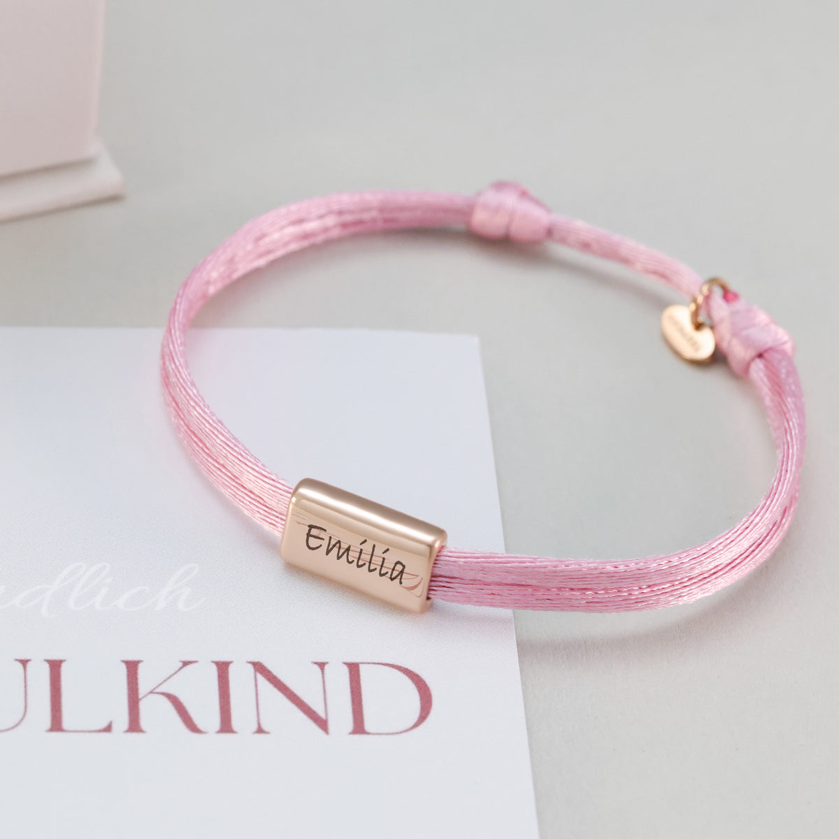 Kinder Satinband Armband mit Gravur in Rosa, Gravur Emilia mit Herzanhänger, auf Einschulungskarte drapiert