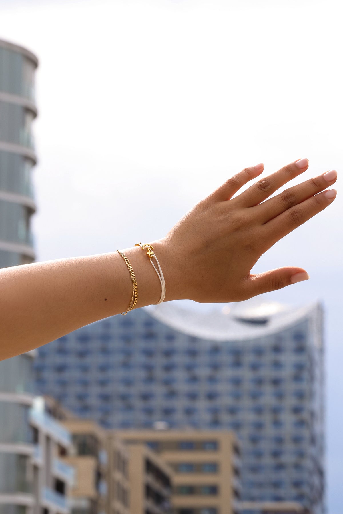 Frau trägt weißes Unisex Armband mit Gravur & Ankerhaken kombiniert mit Goldkette, Arm vor Hochhaus-Kulisse