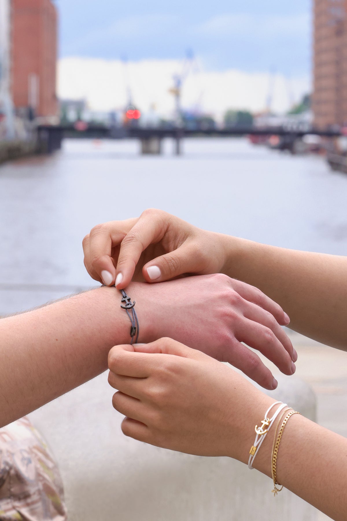 Zwei Personen befestigen graues Ankerhaken Nylon Armband mit Gravur am Handgelenk vor Hafenkulisse