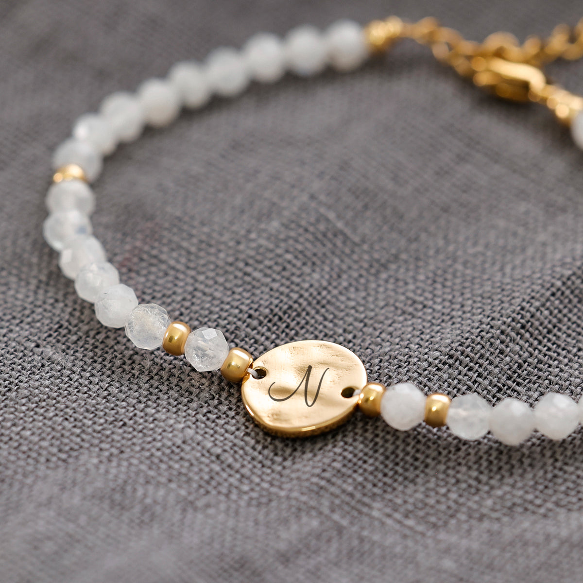 Mondstein Armband mit Hammerschlag Plättchen in Gold, Nahaufnahme mit Initiale auf Stoffuntergrund